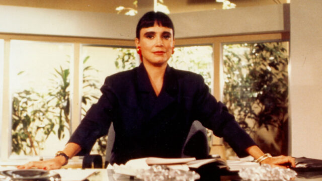 Regina Duarte como a protagonista Maria do Carmo com blazer escuro e cabelo curto, sentada em seu escritório em cena de Rainha da Sucata