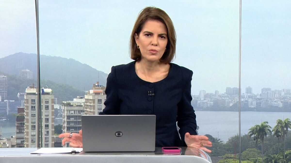 A jornalista Mariana Gross com blazer escuro comanda o telejornal RJ1 na bancada da Globo com vista para o Rio de Janeiro