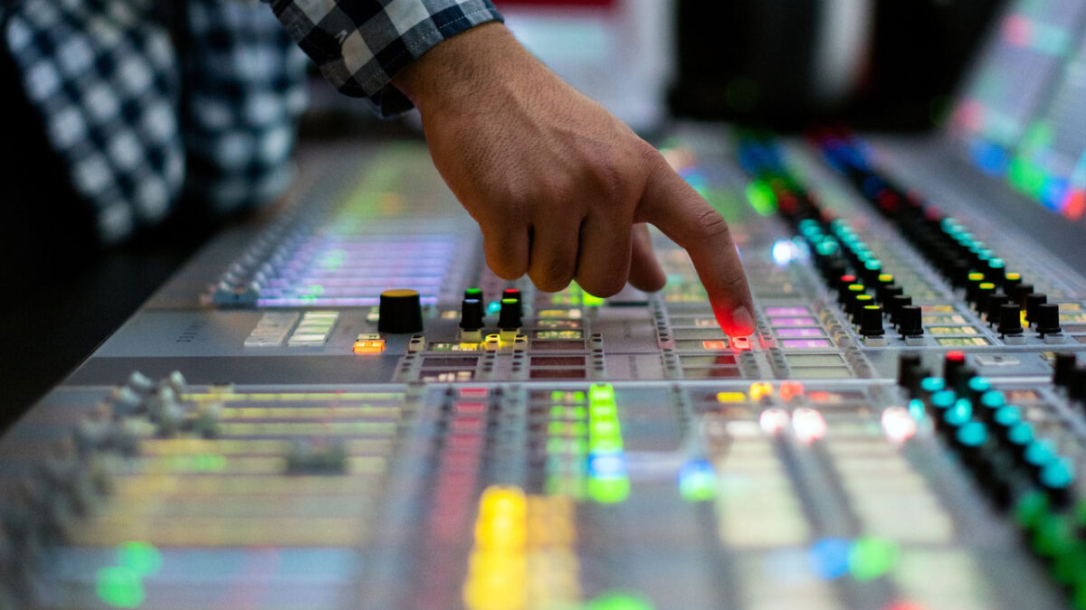 Mão de um operador pressiona um botão iluminado em uma mesa de som ou transmissão profissional, com faders e luzes coloridas