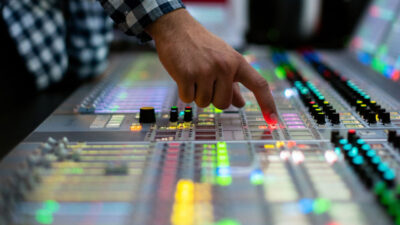 Mão de um operador pressiona um botão iluminado em uma mesa de som ou transmissão profissional, com faders e luzes coloridas