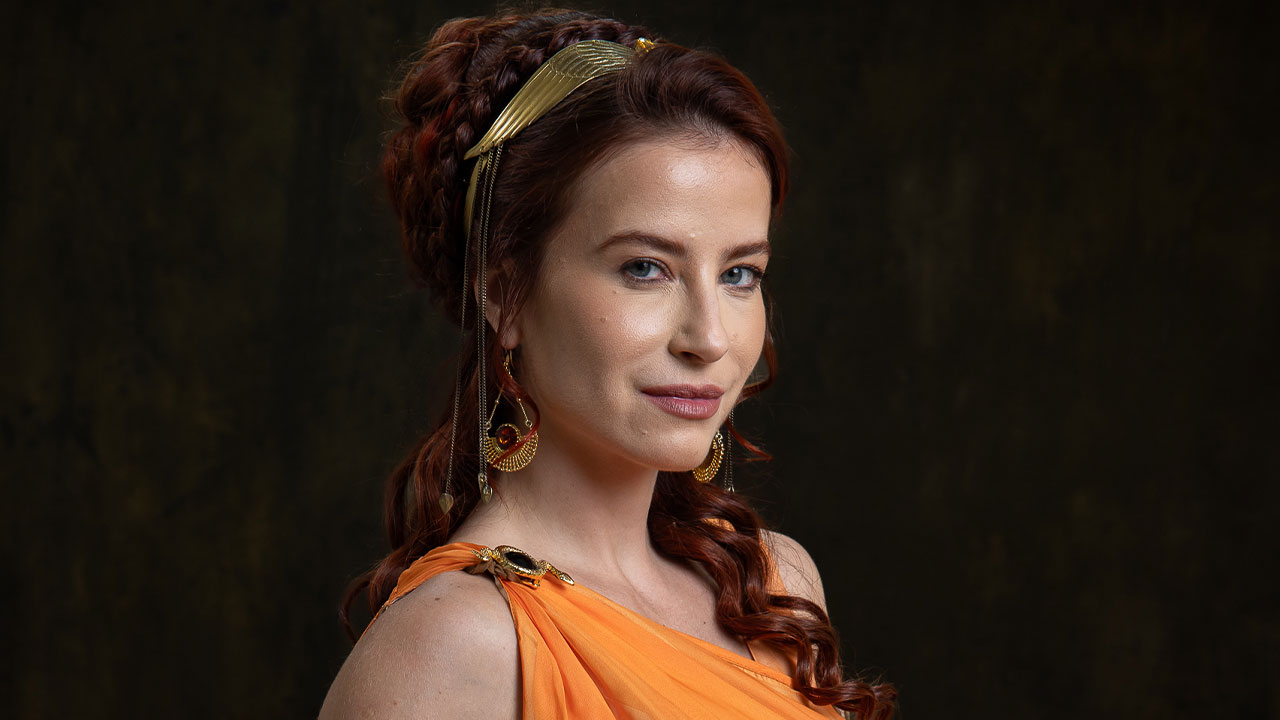 Messalina (Bárbara França) com cabelo ruivo preso, tiara e vestido laranja em foto de divulgação de O Senhor e a Serva