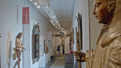 Interior do Museu de Arte Sacra de SP com esculturas do Século XVIII e visitantes no fundo