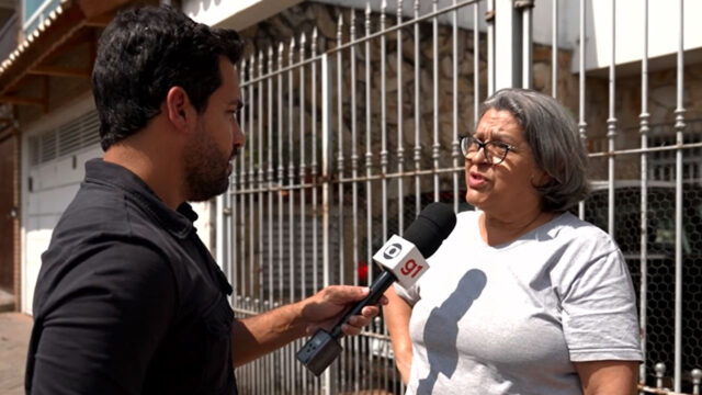 O repórter Philipe Guedes, com camisa preta, entrevista uma mulher de óculos e camisa branca com microfone da Globo na rua