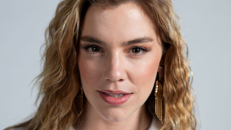 Alice Wegmann, com cabelos loiros e ondulados, caracterizada como a personagem Raíssa Medeiros da série Rensga Hits!