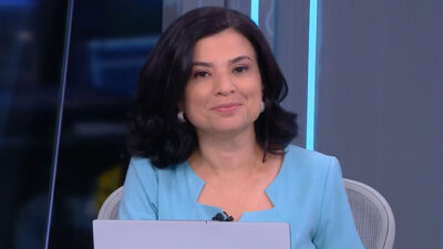 A jornalista Raquel Landim sorri com cabelo escuro e blazer azul claro, sentada em frente a um notebook em estúdio da CNN Brasil