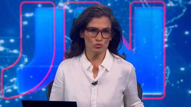 Renata Vasconcellos apresenta o Jornal Nacional usando camisa branca e óculos diante do telão azul com as iniciais JN