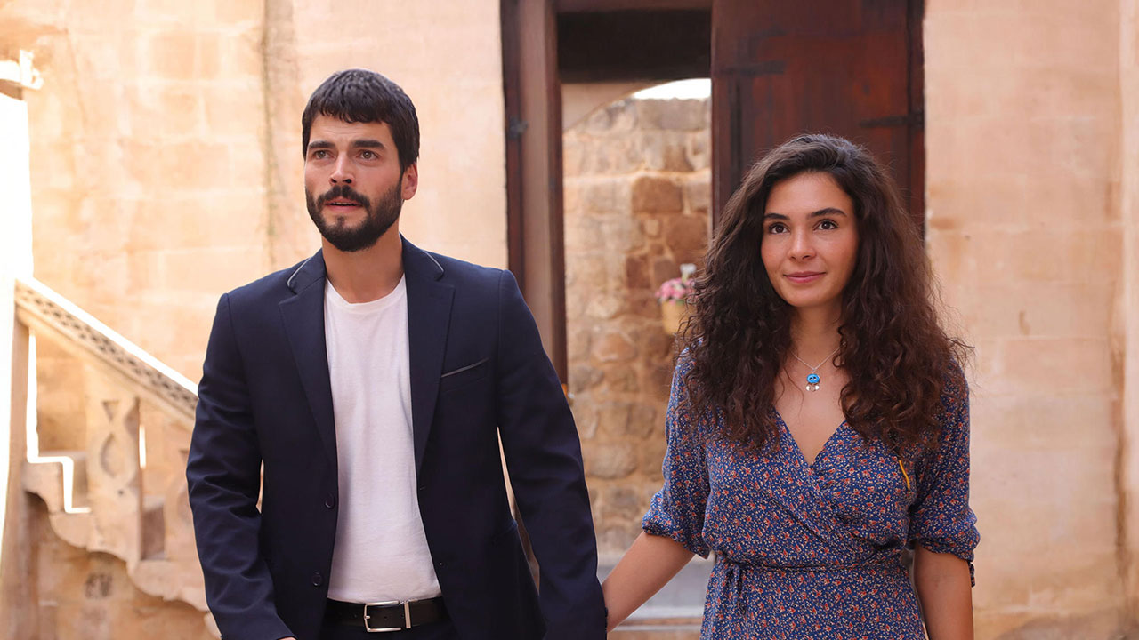 Reyyan (Ebru Sahin) e Miran (Akin Akinözü) de mãos dadas caminhando em pátio de pedra na novela turca Hercai