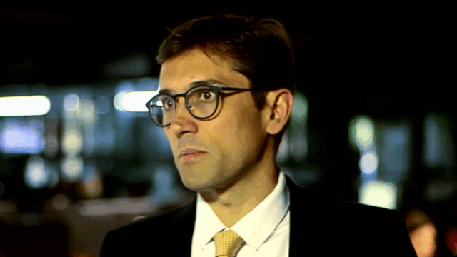 Rodolfo Schneider, diretor de Jornalismo da Band, com óculos e terno preto, olha sério para o lado em gravação na Redação