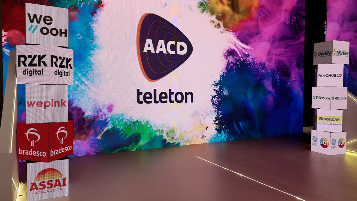 Cenário do Teleton 2025 com o logo da AACD e das empresas patrocinadoras sobre caixas de LED e telão ao fundo com arte em tons coloridos