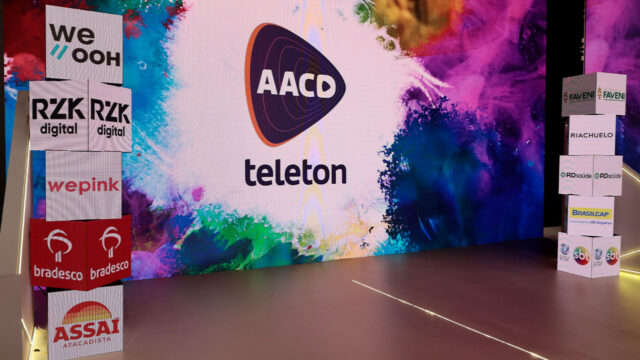 Cenário do Teleton 2025 com o logo da AACD e das empresas patrocinadoras sobre caixas de LED e telão ao fundo com arte em tons coloridos