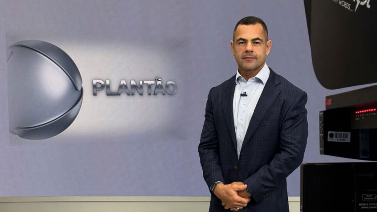 O jornalista Tino Júnior, de terno azul-marinho, apresenta o Plantão da Record em um estúdio com fundo claro