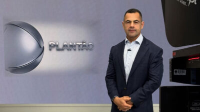 O jornalista Tino Júnior, de terno azul-marinho, apresenta o Plantão da Record em um estúdio com fundo claro