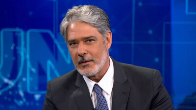 O jornalista William Bonner no Jornal Nacional com terno escuro e gravata, olha sério no estúdio com fundo azul