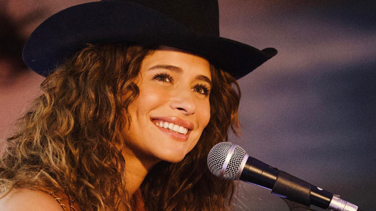 Isadora Cruz como Agrado Garcia sorri com chapéu de cowboy e cabelo cacheado cantando em um microfone na novela Coração Acelerado