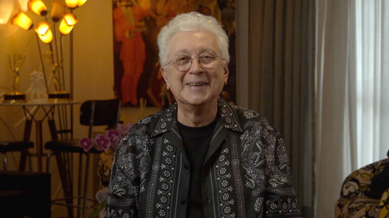 O autor de novelas Aguinaldo Silva sorrindo, de óculos e camisa preta estampada, em ambiente interno com luz quente