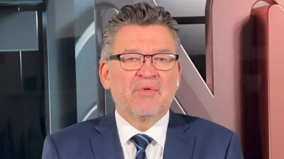 Américo Martins, jornalista com óculos de aro preto, usando terno e gravata, no estúdio da CNN Brasil com iluminação em tons de cobre