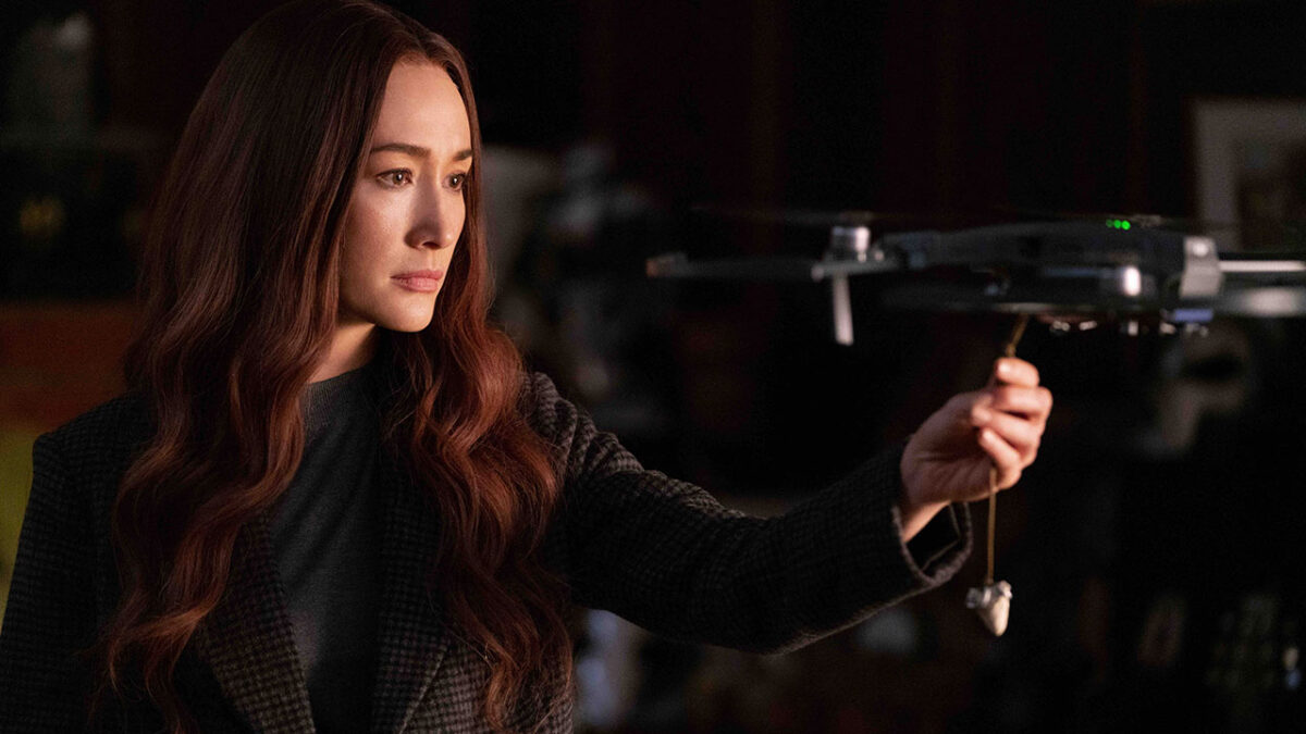 Maggie Q como Anna, com cabelo comprido e jaqueta escura, segurando um drone em cena de ação do filme A Profissional