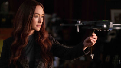 Maggie Q como Anna, com cabelo comprido e jaqueta escura, segurando um drone em cena de ação do filme A Profissional