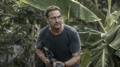 Gerard Butler como Brodie Torrance, com uma camiseta escura e ferimentos no rosto, segurando uma arma em uma selva