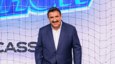 O apresentador Carlos Massa, o Ratinho, sorri com terno azul-marinho e camisa branca, em frente a um cenário com uma rede de futebol