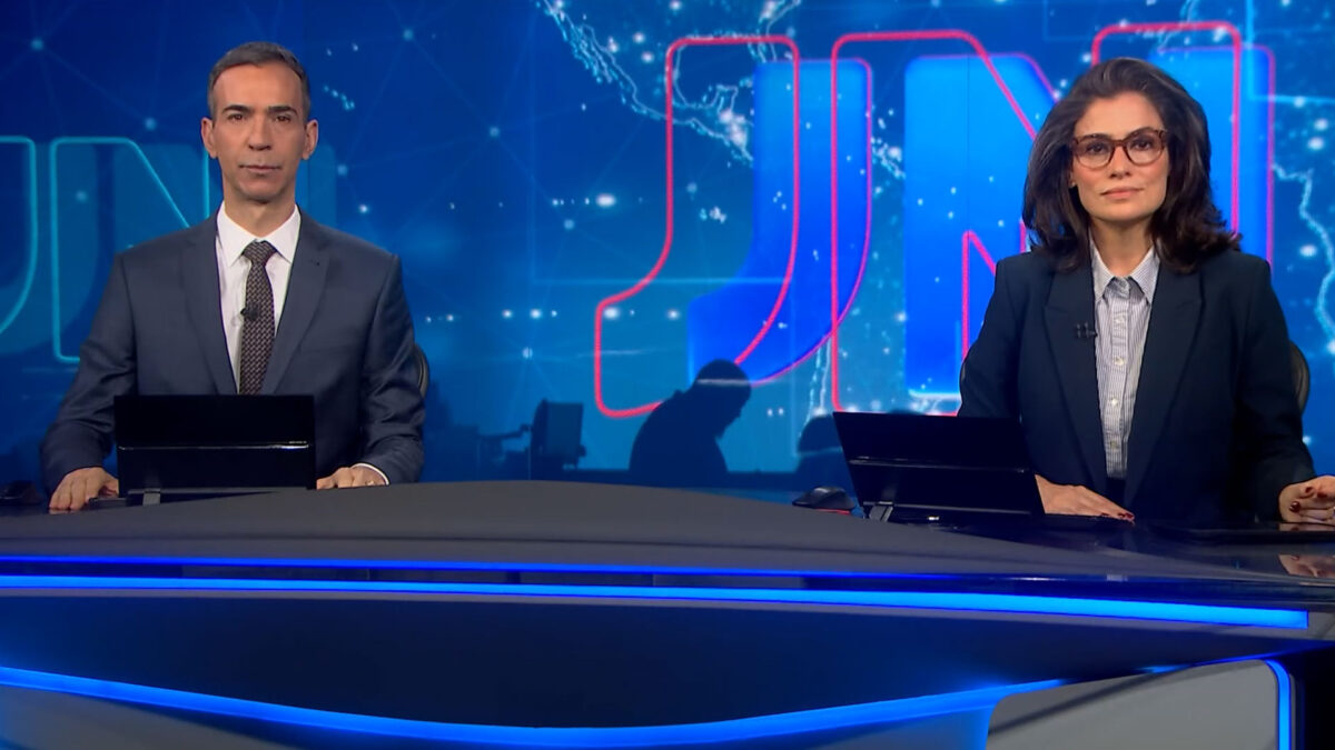 César Tralli e Renata Vasconcellos na bancada do Jornal Nacional, ambos de terno, com o telão do JN em azul ao fundo