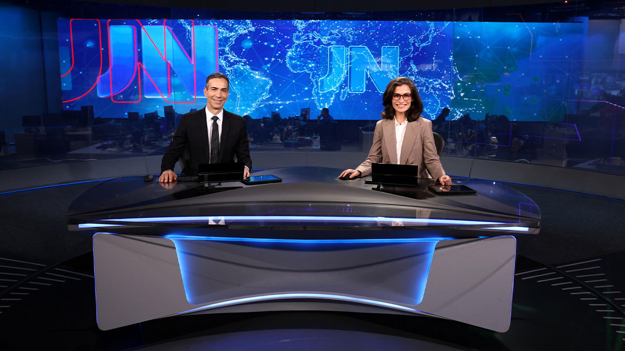 César Tralli, de terno escuro, e Renata Vasconcellos, de blazer bege, sorriem na bancada do Jornal Nacional com o telão do JN ao fundo