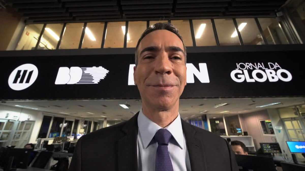 O jornalista César Tralli sorri na redação da Globo com o logo dos telejornais de rede em destaque na parede ao fundo