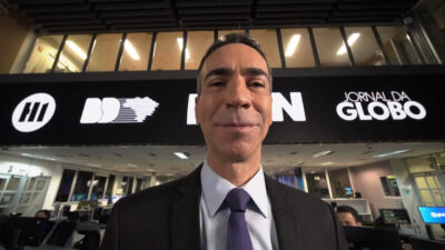 O jornalista César Tralli sorri na redação da Globo com o logo dos telejornais de rede em destaque na parede ao fundo