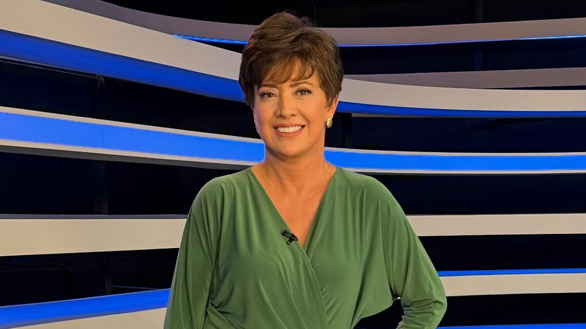 A jornalista Christina Lemos sorri com blusa verde e cabelo curto, em pé no estúdio do Jornal da Record com listras azuis ao fundo