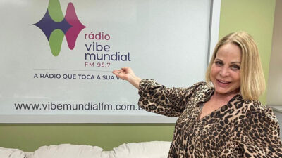 Claudete Troiano, sorrindo e de braços estendidos, usando um vestido de oncinha em frente ao logotipo da Rádio Vibe Mundial