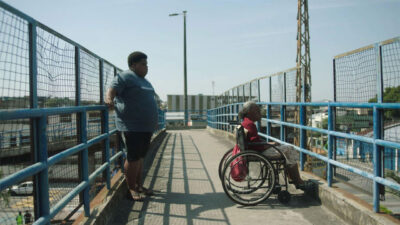 Dé (Big Jaum) e Dona Almerinda (Teca Pereira) em cadeira de rodas, parados no alto de uma passarela de metal em cena do filme Kasa Branca