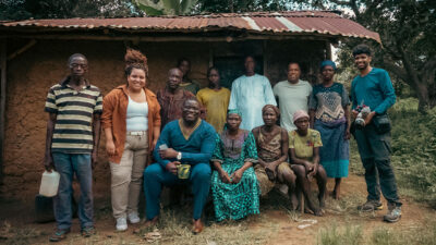 Equipe do Diários da Nigéria da GloboNews e moradores locais em frente a uma casa simples de telhado de zinco na África