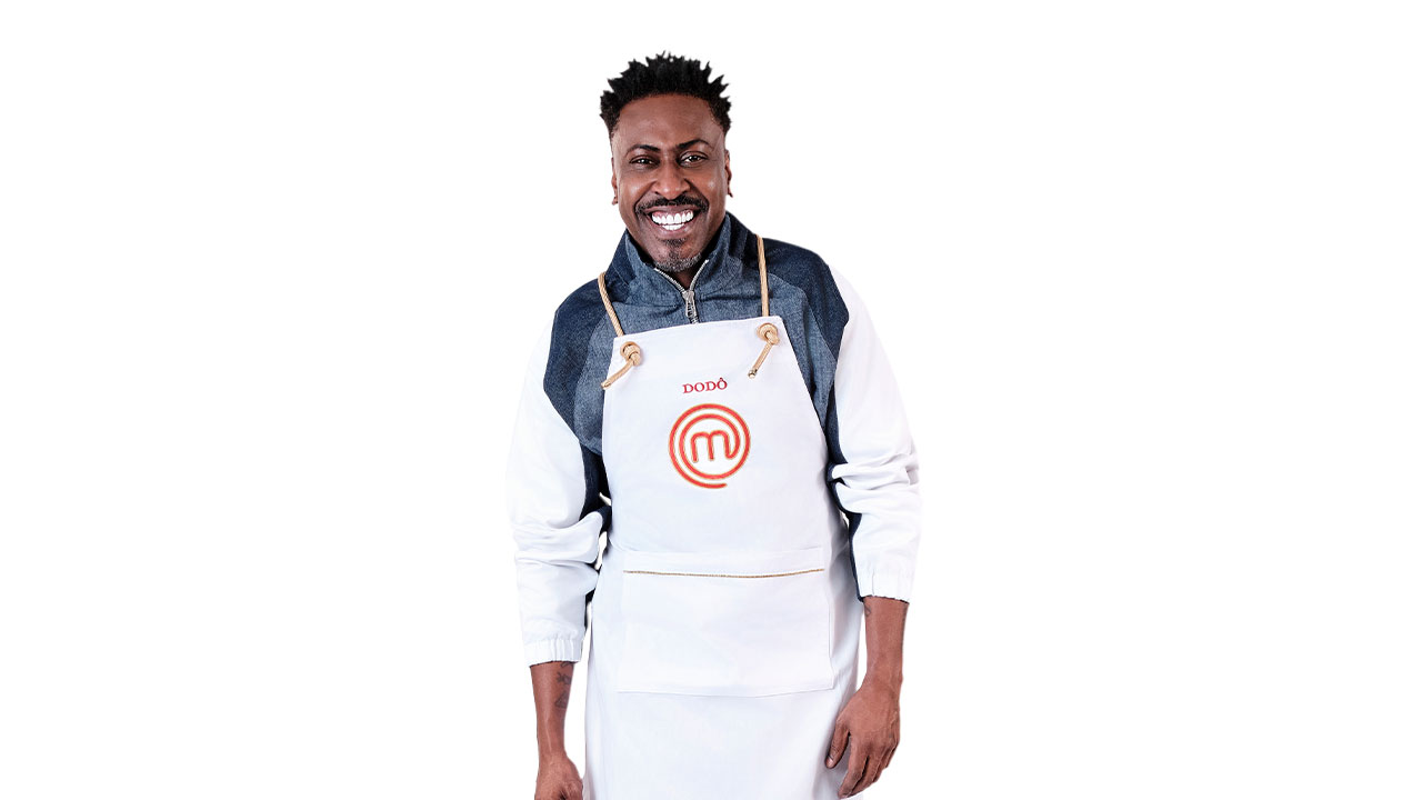 Dodô, do grupo Pixote, sorrindo, usando um casaco cinza e branco sobre o avental branco MasterChef com seu nome