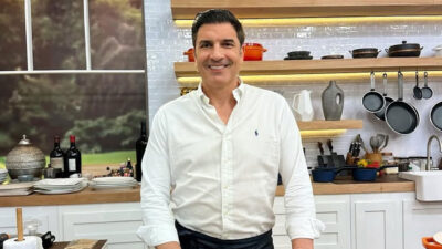 Chef e apresentador Edu Guedes sorrindo, usando camisa branca em pé na cozinha decorada do programa Fica Com a Gente