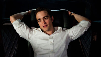 Ator Robert Pattinson como Eric Packer, com camisa branca, deitado no banco de trás de uma limusine em Cosmópolis