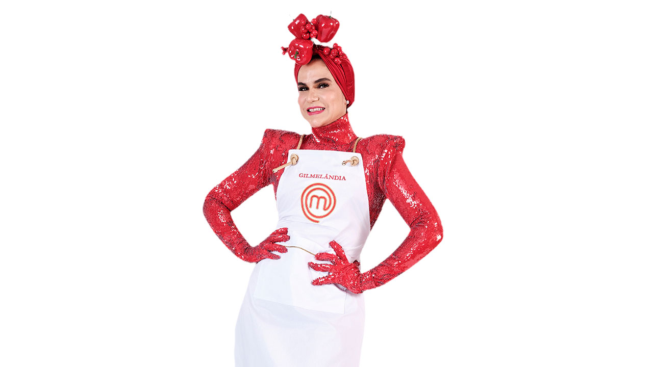 Gilmelândia com roupa e turbante vermelho brilhante, sorrindo e posando com avental branco MasterChef para o programa de culinária