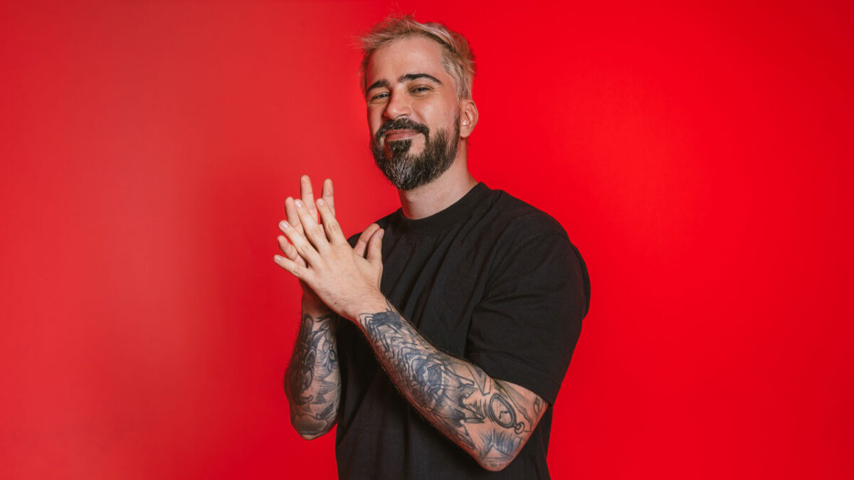 O apresentrador Guilherme Guedes, com barba e cabelo loiros, sorri para a câmera batendo as mãos em fundo vermelho