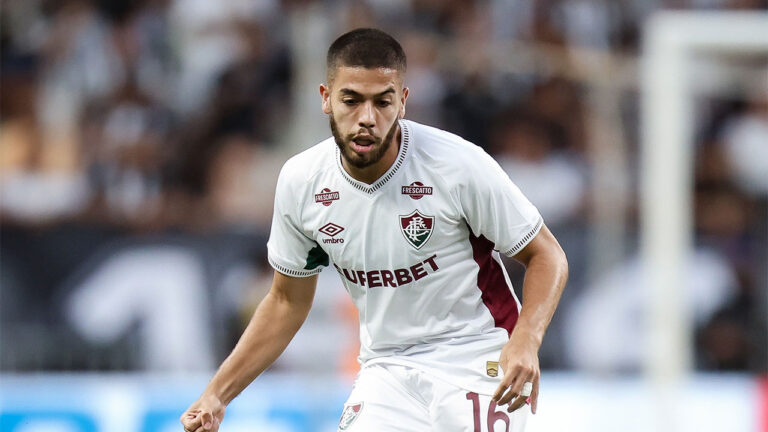 O jogador Gustavo Nonato com a camisa branca e grená do Fluminense, no campo, olha fixamente para a partida de futebol