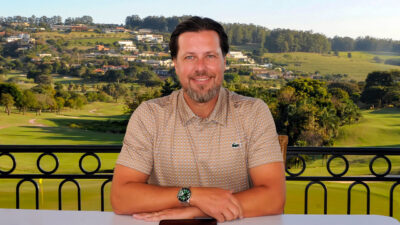 Henrique Kirilauskas usando camisa polo estampada e relógio, sorrindo em uma varanda com vista para um campo de golfe