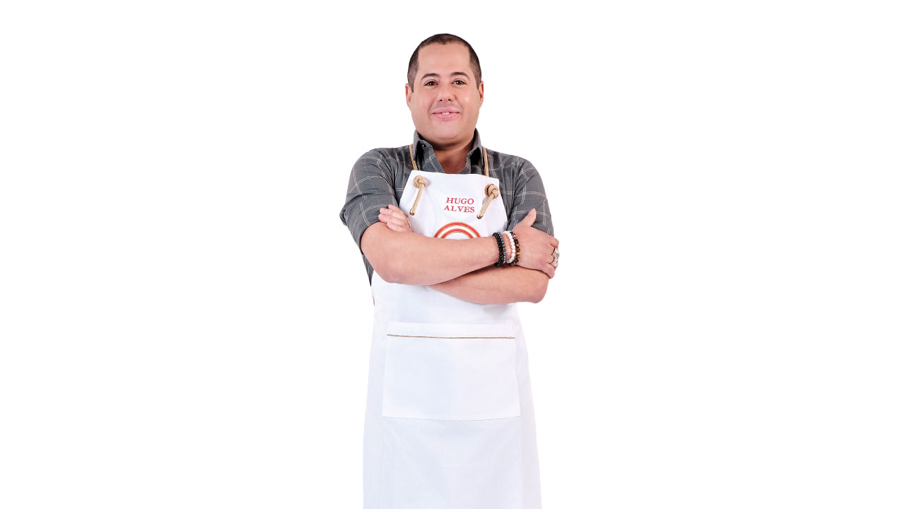 Hugo Alves sorrindo para a câmera com os braços cruzados, usando uma camisa xadrez escura e o avental branco MasterChef