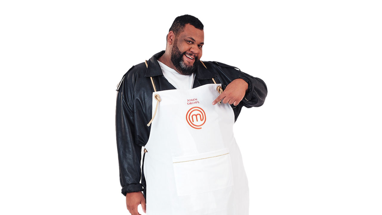 John Drops com barba, apontando para o avental branco do MasterChef com seu nome, usando uma jaqueta de couro preta