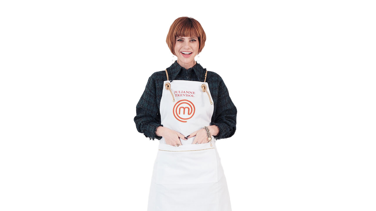 Julianne Trevisol sorrindo de cabelo ruivo curto com franja, vestindo avental branco MasterChef sobre blusa escura e as mãos no bolso