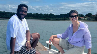 Ator Lázaro Ramos e Sandra Annenberg de óculos escuros e casaco lilás sorrindo em um barco no lago ou mar