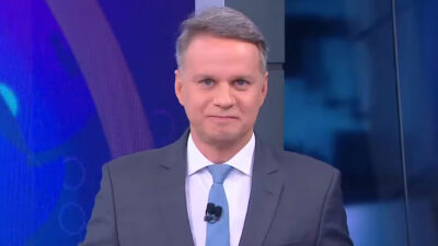 O jornalista Leandro Magalhães, de terno cinza e gravata azul, apresenta telejornal com olhar sereno em estúdio com fundo azul