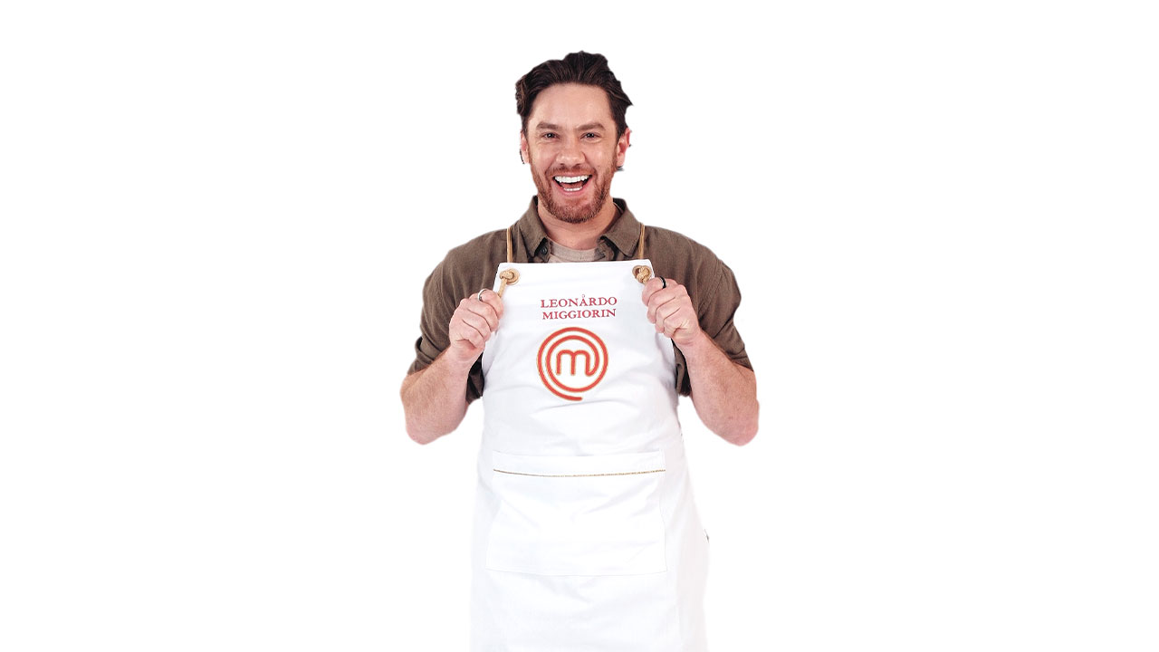 Leonardo Miggiorin sorridente, puxando o avental branco MasterChef com o nome dele e barba em foto de divulgação do reality