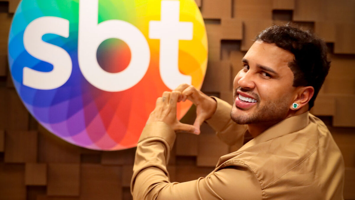 Apresentador Lucas Guimarães sorrindo e fazendo um coração com as mãos em frente ao logotipo colorido do SBT na parede