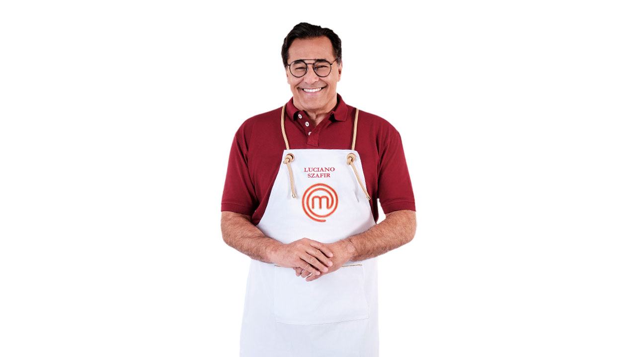 Luciano Szafir sorrindo, usando óculos, camisa polo vermelha e o avental branco do MasterChef, com as mãos cruzadas