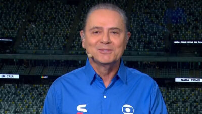 Narrador Luís Roberto sorrindo, usando camisa social azul em frente à arquibancada escura de um estádio de futebol