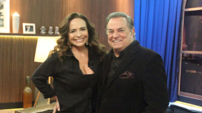 Luíza Tomé de blusa preta, sorrindo ao lado de Ronnie Von de blazer preto, no cenário do programa Companhia Certa