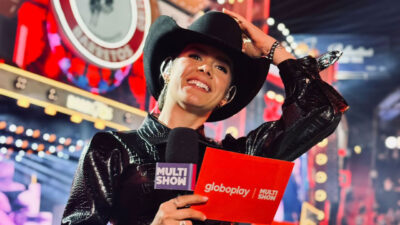Marina Fabris sorrindo e usando chapéu de cowboy preto, segurando microfone e uma ficha de apresentador com logos do Globoplay e Multishow